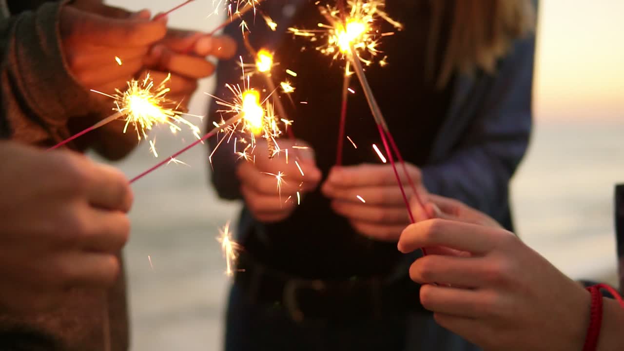 Close Up view of hands of group of people holding bengal lights and lightning them up on the beach during sunset