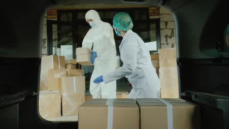 Movers-In-Protective-Suits-Load-Cardboard-Boxes-With-Medicine-Into-The-Car