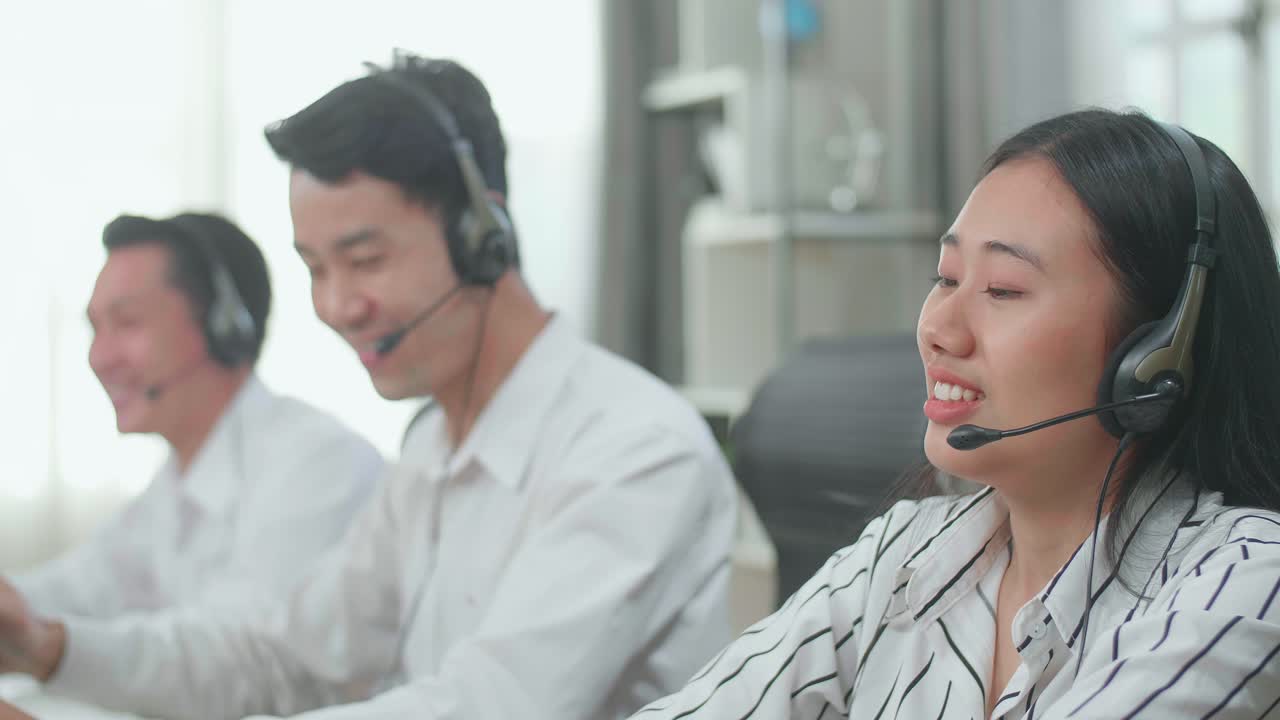 Three Asian Call Center Agents Wearing Headsets Sitting On The Chairs And Being Happy Due To Success Working With Computers At The Office