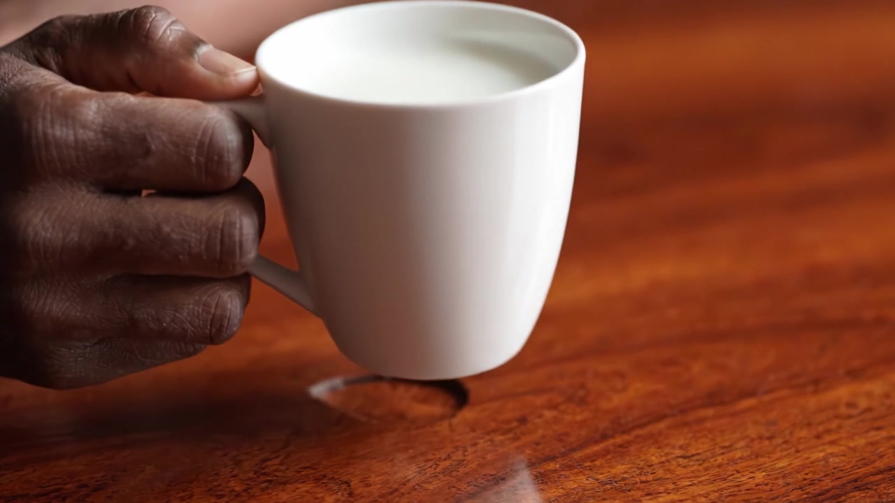 Una mano che tiene una tazza bianca di latte su una superficie di tavolo in legno, catturando un momento di serenità e comfort in un ambiente accogliente
