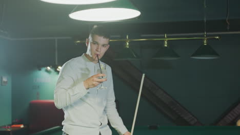 player in white shirt holds cue stick and sips lemon drink with black straw while pacing beside green pool table, observing ball placement under focused lighting in a modern billiards lounge