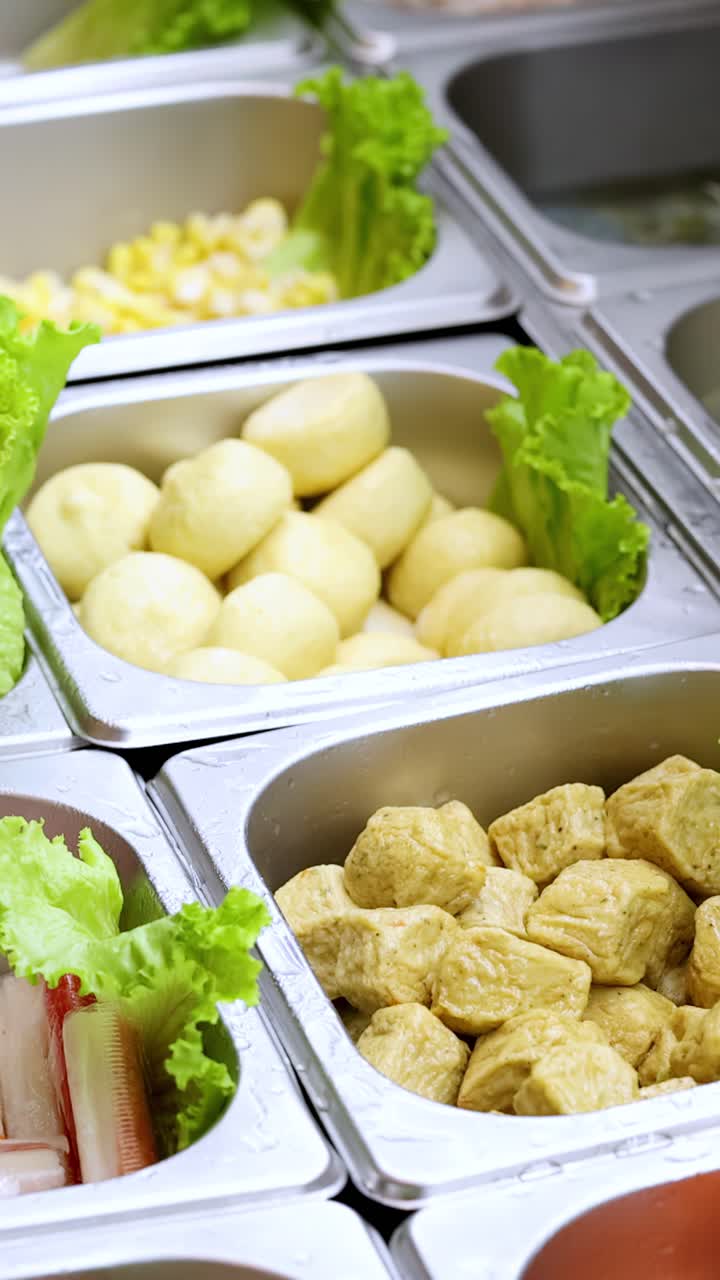 Panoramica verticale su vassoi da buffet in acciaio inossidabile con ingredienti assortiti per hotpot sotto una luminosa illuminazione interna.