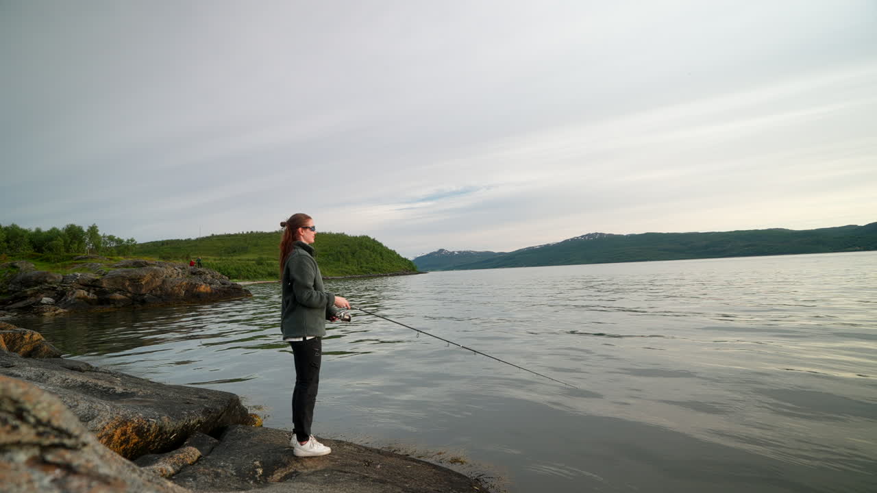 Woman casting a fishing rod from rocky shore in Tromsø, Norway, surrounded by calm fjord waters and scenic wilderness under an overcast Arctic sky