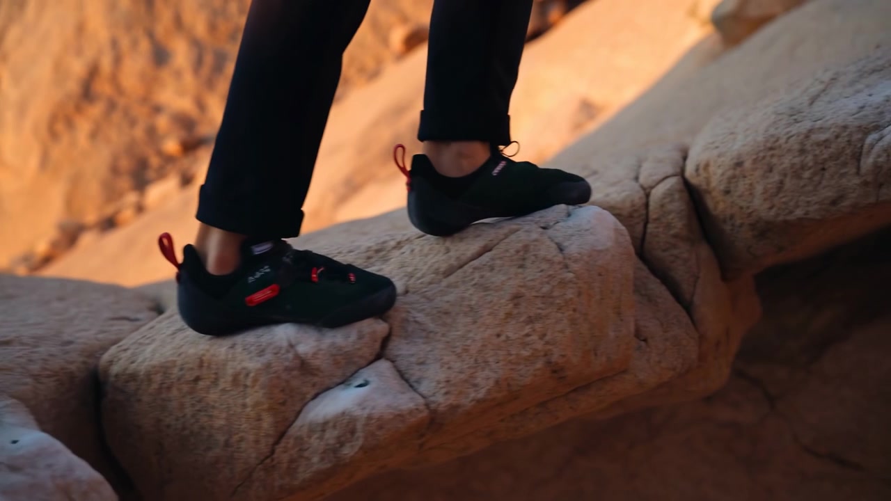 Lower legs and feet wearing black climbing shoes with red pull tabs step along narrow sandstone ledge, visible motion shows rear foot lifting, front foot pressing, and careful balance in warm canyon