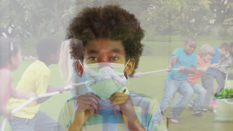 african american boy wearing a face mask against group of diverse kids playing tug of war in garden