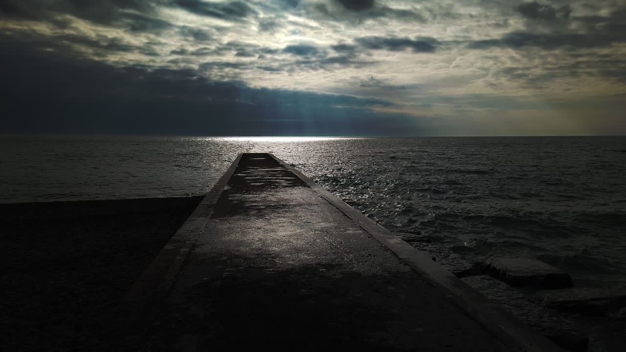A long wet concrete jetty extending from the shoreline out to Lake Ontario. wide shot