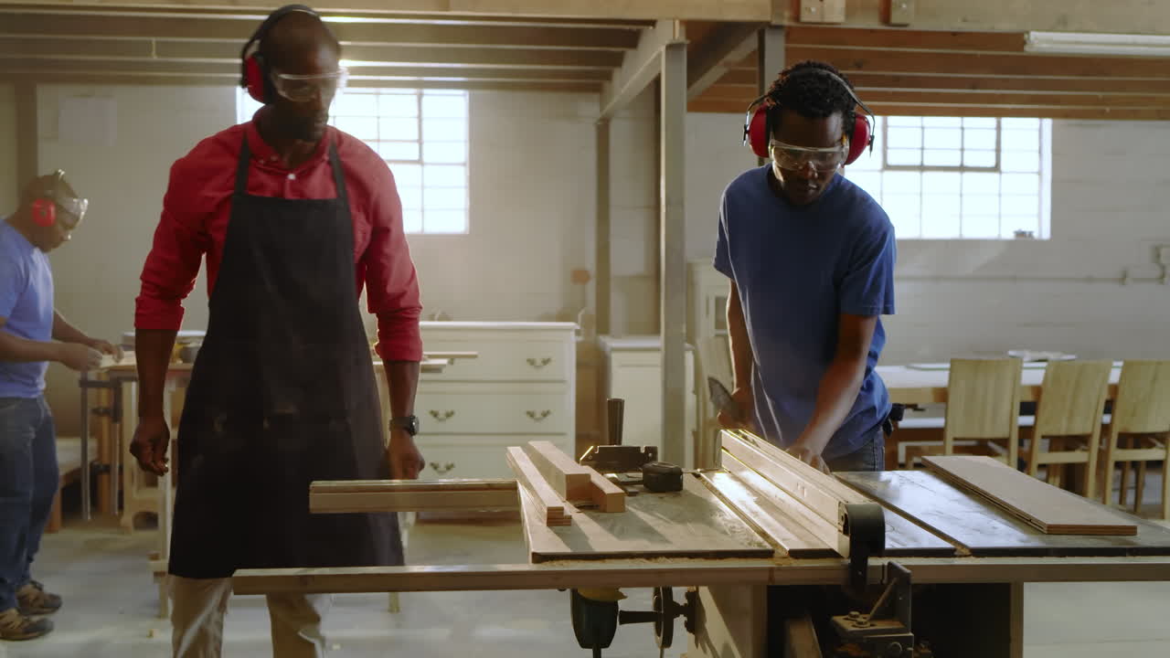 Mid adult African American man with goggles feeding plank at table saw after colleague marking wood