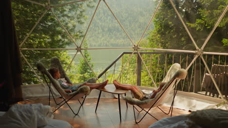 children rest in glamping cabin at lakeside. little boy and girl sit in comfortable chairs under transparent dome. kids enjoy lake view at camp