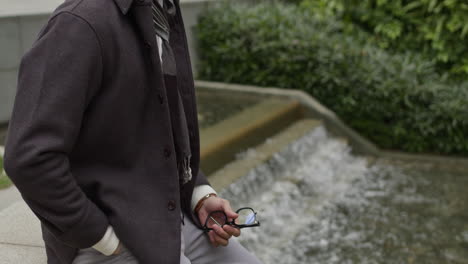 caucasian modern male with trendy clothes, man model sitting in front of a water river holding his glasses in his hands while thinking