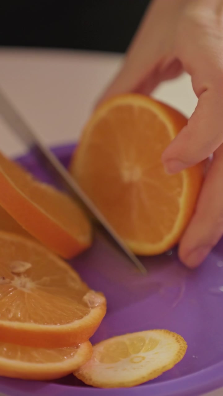 Primo piano di una mano che affetta un'arancia su un piatto viola. Le fette d'arancia si accumulano, con una fetta di limone visibile tra di esse. Ambientazione interna con sfondo neutro e messa a fuoco ravvicinata stabile.