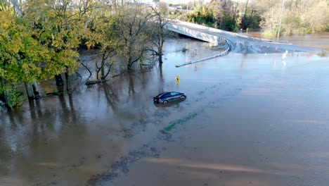 Überflutetes-Blaues-Auto,-Das-In-Einem-Angeschwollenen-Fluss-Gestrandet-Ist,-Der-Straße-Und-Brücke-Bedeckt,-Luftaufnahme