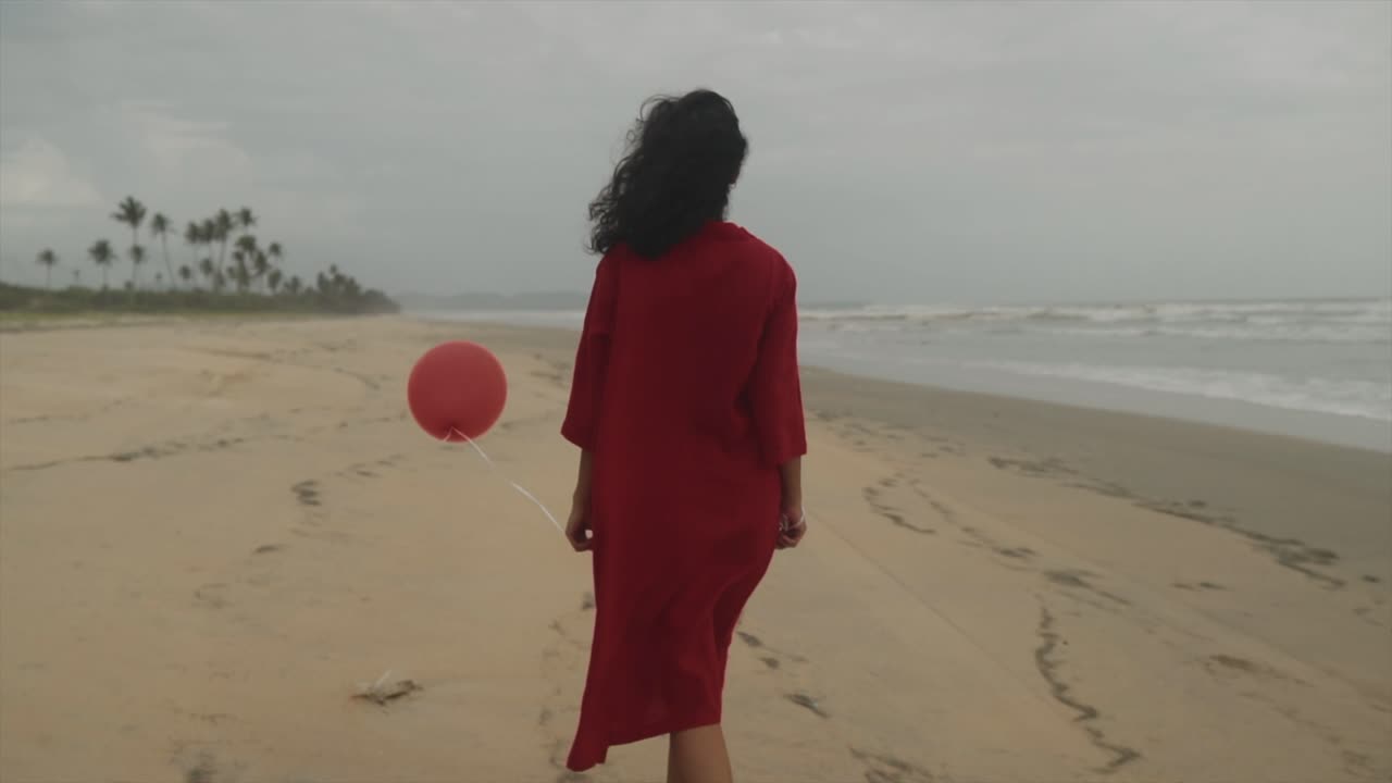 Bela jovem indiana morena com um vestido vermelho e um balão vermelho na mão a caminhar pela praia do Mar Arábico em Goa, Índia, num dia nublado
