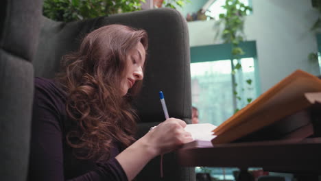 side view of woman with long wavy brown hair writing attentively in notebook beside window, surrounded by soft indoor lighting and lush blurred greenery in background, peaceful learning atmosphere