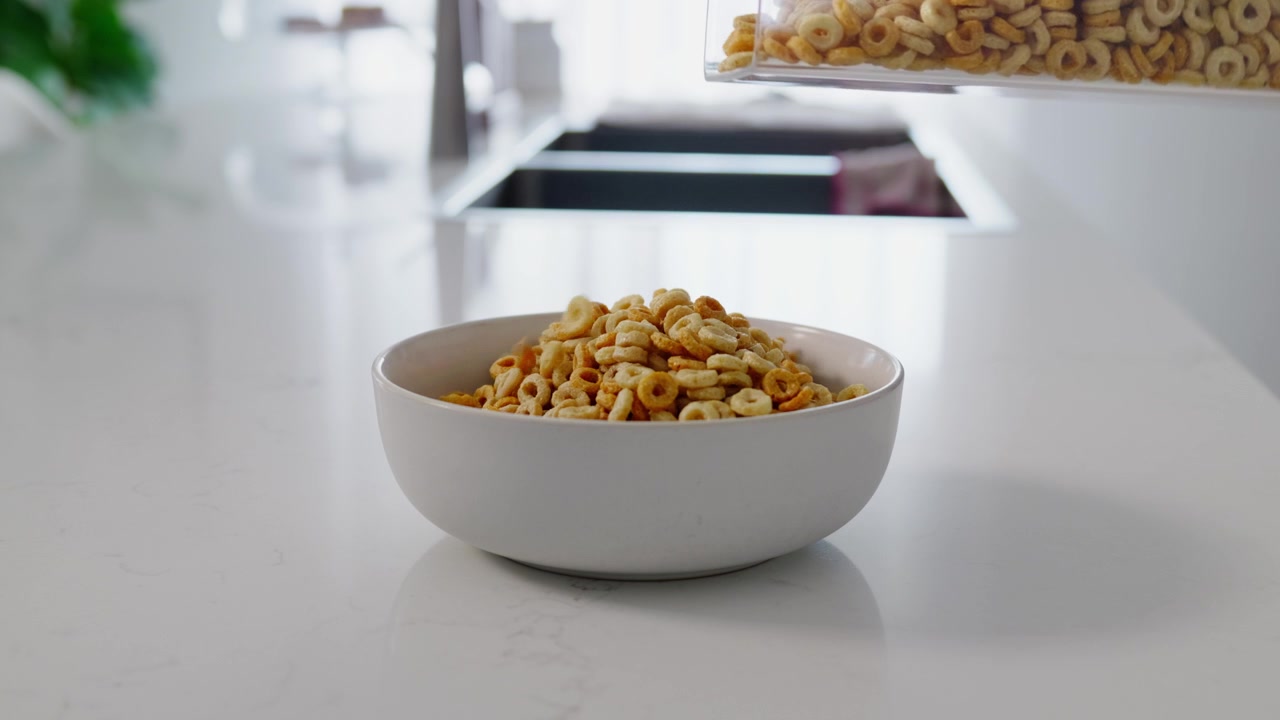 Primo piano di cereali per la colazione versati in una ciotola su un bancone della cucina. Fiocchi croccanti cadono nella ciotola, catturando una semplice routine mattutina e una scena di preparazione del cibo.