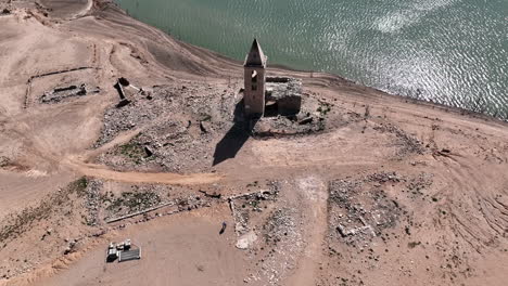 Aerial-view-rising-over-abandoned-Church-of-Sant-Roma,-Catalan-tower-on-deserted-rocky-coastline-of-the-Sau-reservoir