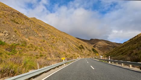 a vehicle travels down a winding rural road bordered by grassy hills and mountains under partly cloudy skies, captured with smooth forward camera movement and natural lighting