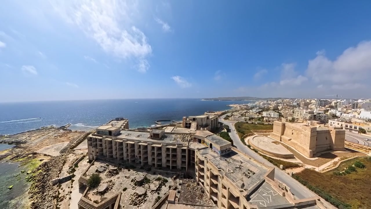 Drone che vola all'indietro sopra l'hotel resort abbandonato e in rovina a Marsaskala, Malta, con la torre di avvistamento di San Tumas sullo sfondo.