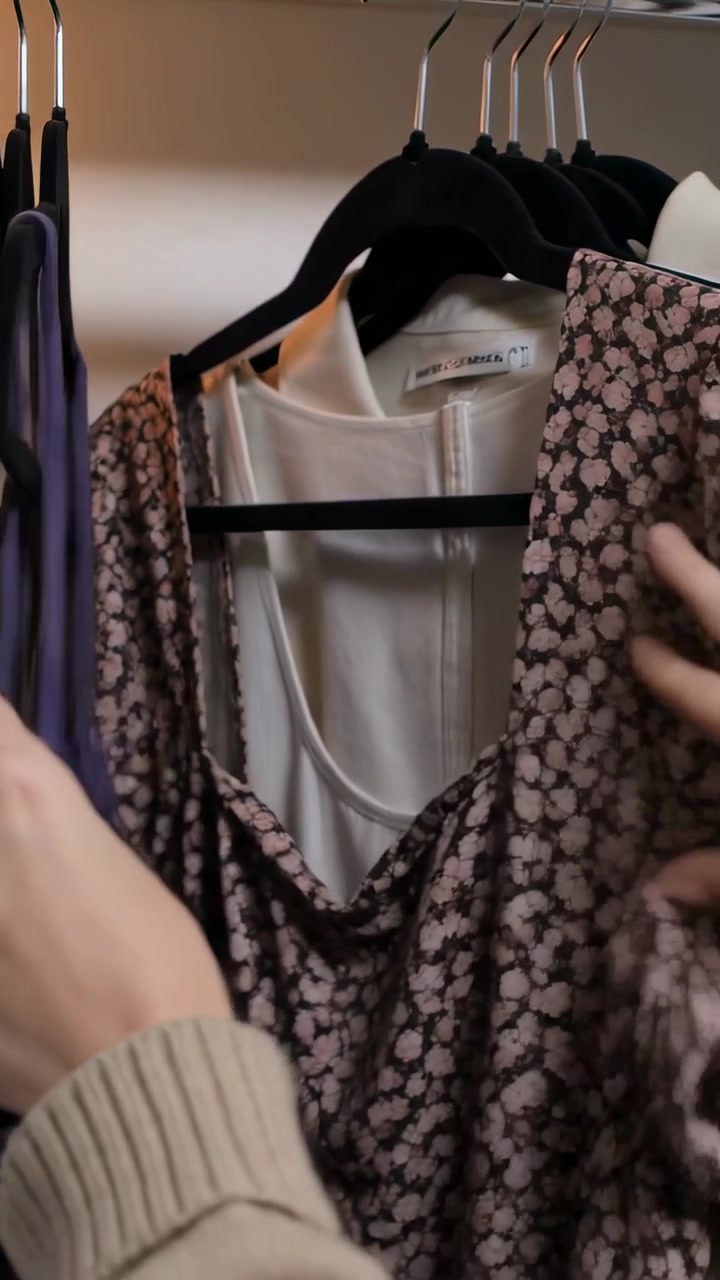 Video verticale: Mani con manica color cuoio che si allungano, tirano un vestito floreale, scegliendo una camicetta bianca nell'armadio