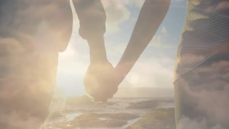 animation of blue sky and clouds over biracial couple holding hands at beach