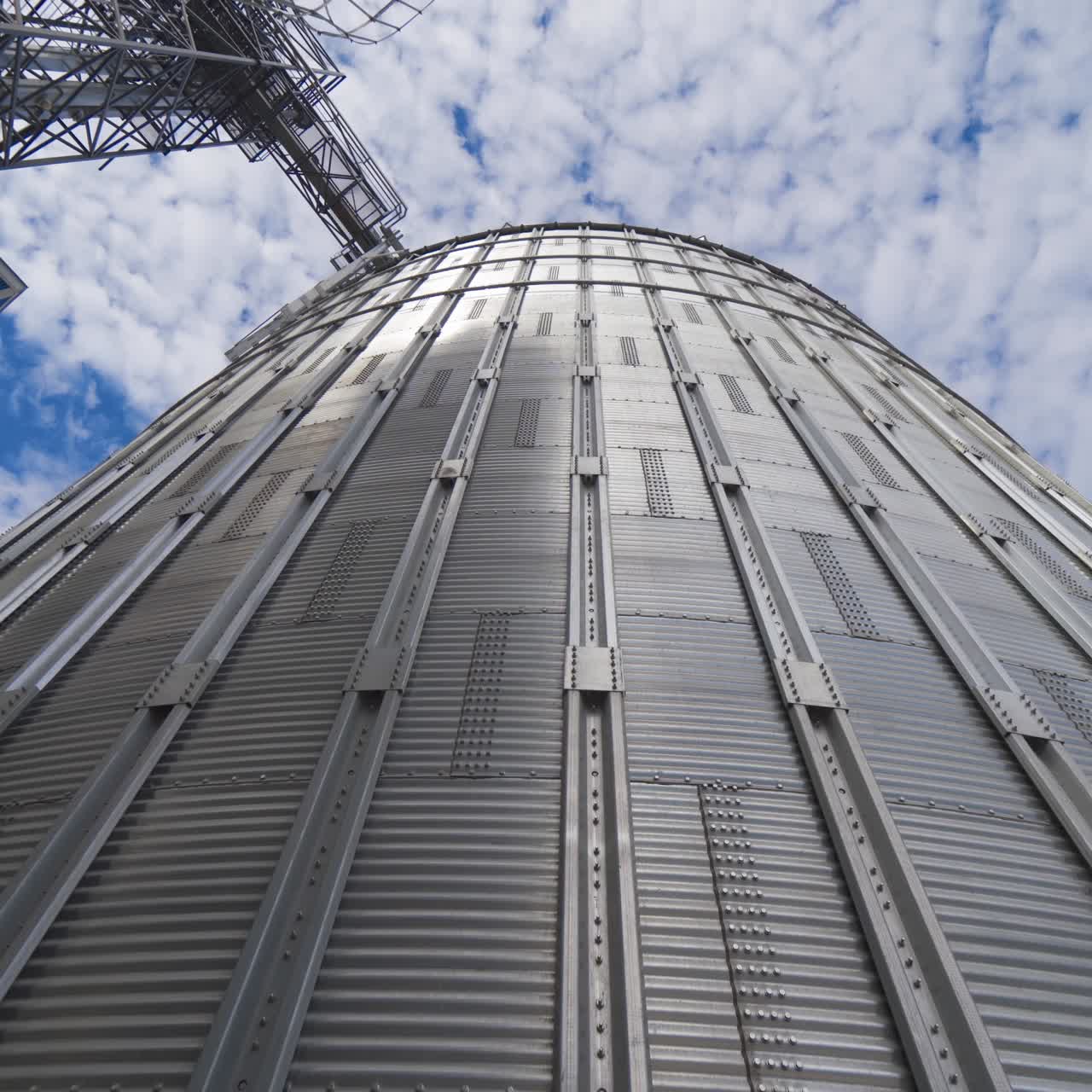 Large metal grain elevator outdoors. Modern silver granary on sky background. Huge aluminum container for storing crop. View from below