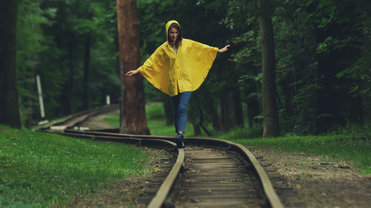 Attractive Girl In A Yellow Raincoat Walking Carefully On The Old Railway In The Forest
