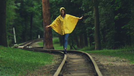 Chica-Atractiva-Con-Un-Impermeable-Amarillo-Caminando-Con-Cuidado-Por-El-Antiguo-Ferrocarril-En-El-Bosque