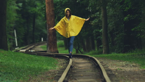 attractive girl in a yellow raincoat walking carefully on the old railway in the forest