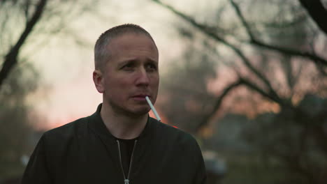 a man in a green jacket standing outdoors, holding a cigarette his mouth, with blurred background with trees and a car