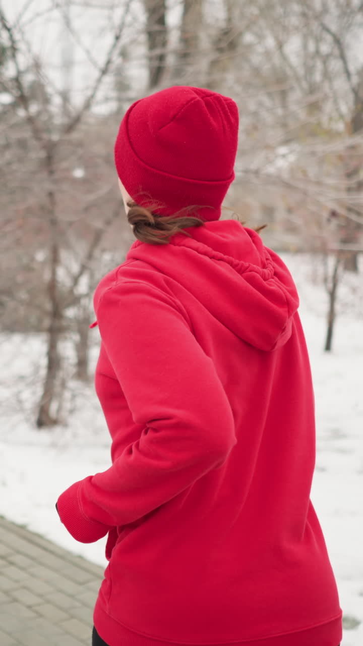 back view of woman jogging along snow covered park pathway flanked by evergreen pines and snow laden trees under foggy winter sky with distant residential building hinting at suburban setting