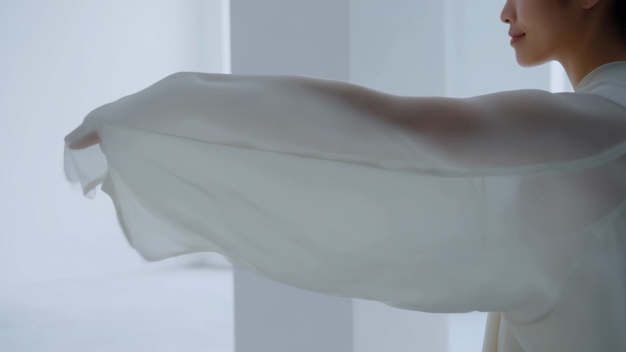 Sidebilde viser en kvinne som strekker ut armene mens hun holder et tynt gjennomsiktig stoff som sprer seg ut i et bredt panel med myke folder, lagdelt gjennomsiktighet og grasiøs bevegelse i et lyst minimalistisk interiør.