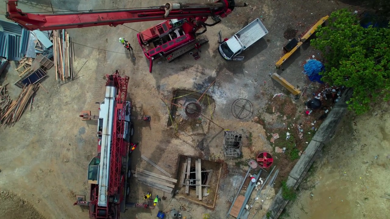 Vista aérea de drone de cima para baixo em ângulo elevado, subindo rapidamente e recuando sobre um poço de fundação de estaca de concreto, máquinas pesadas e trabalhadores em um canteiro de obras de engenharia civil