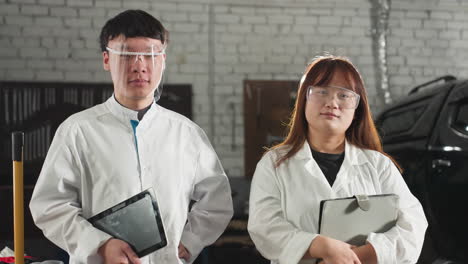 male technician nods with a warm smile while holding a tablet, as his colleague adjusts her glasses with a soft smile, both wear lab coats and stand confidently in an automotive workshop setting