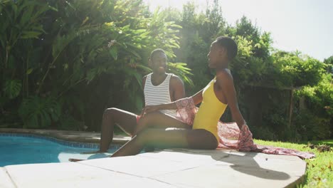 happy african american couple sitting by swimming pool in sunny garden smiling