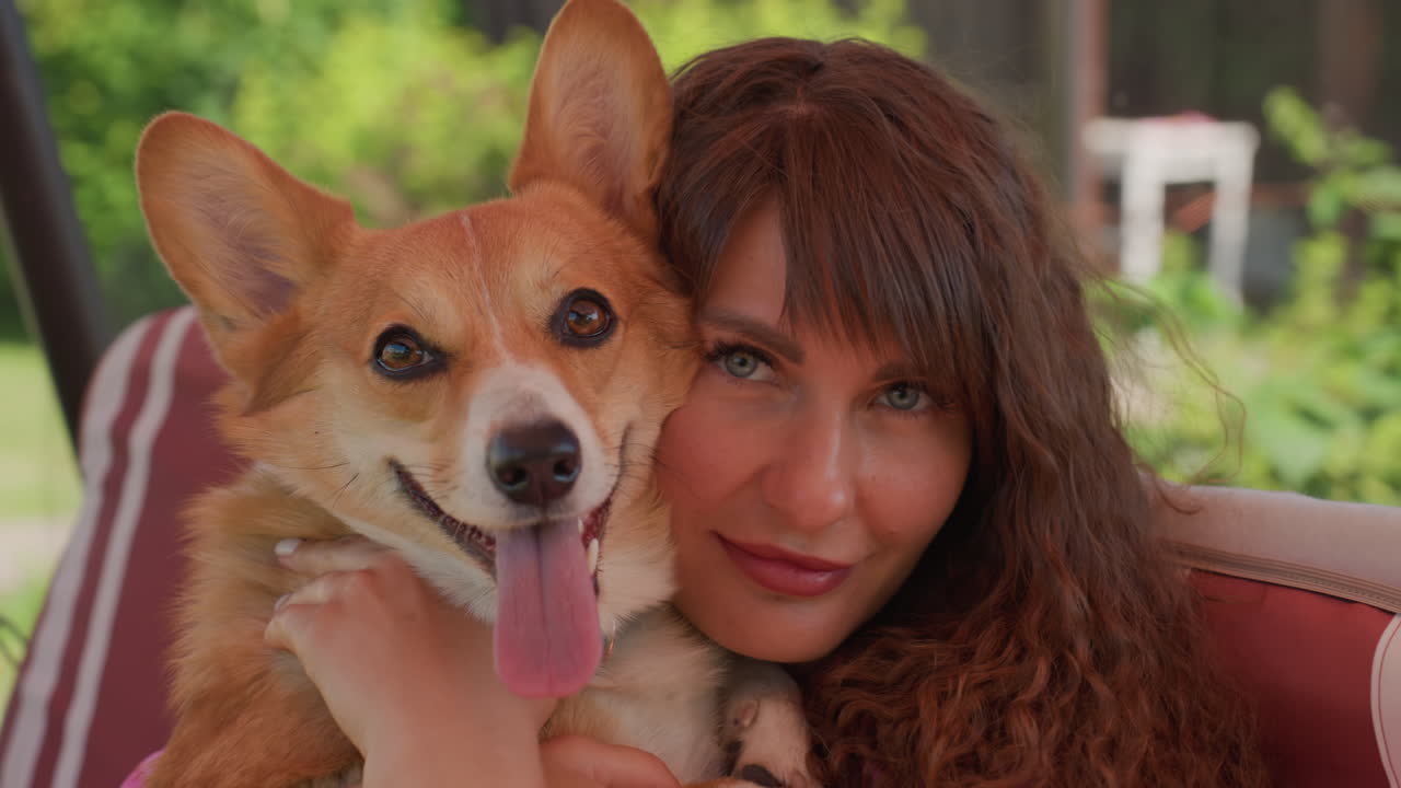 White Mother Hugging Corgi On Striped Swing Sofa, Young Daughter Watching, Sunny Garden Background, Playful TongueOut Dog, CloseUp Faces, Affectionate Family Bond, Casual Summer Clothing, Warm Color