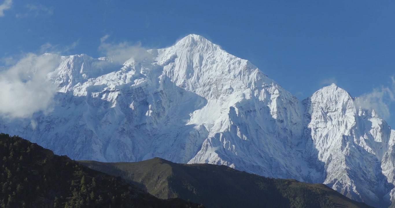 Una ripresa aerea fluida e orbitante circonda il Monte Nilgiri nel Mustang, Nepal, catturando l'imponente vetta himalayana coperta di neve a pieno formato sotto un cielo limpido e soleggiato punteggiato da soffici nuvole bianche.