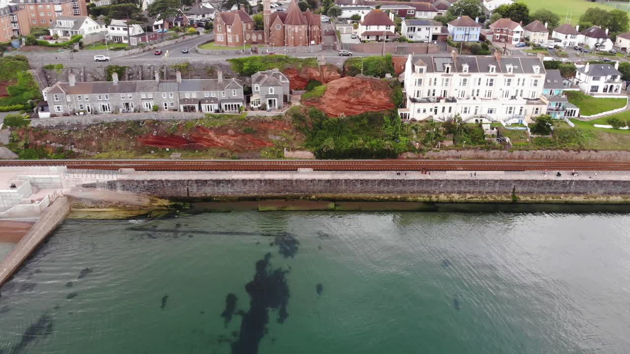 Aerial view of the Dawlish sea wall running alongside the railway line, with the town and residential buildings visible in the background. dolly right