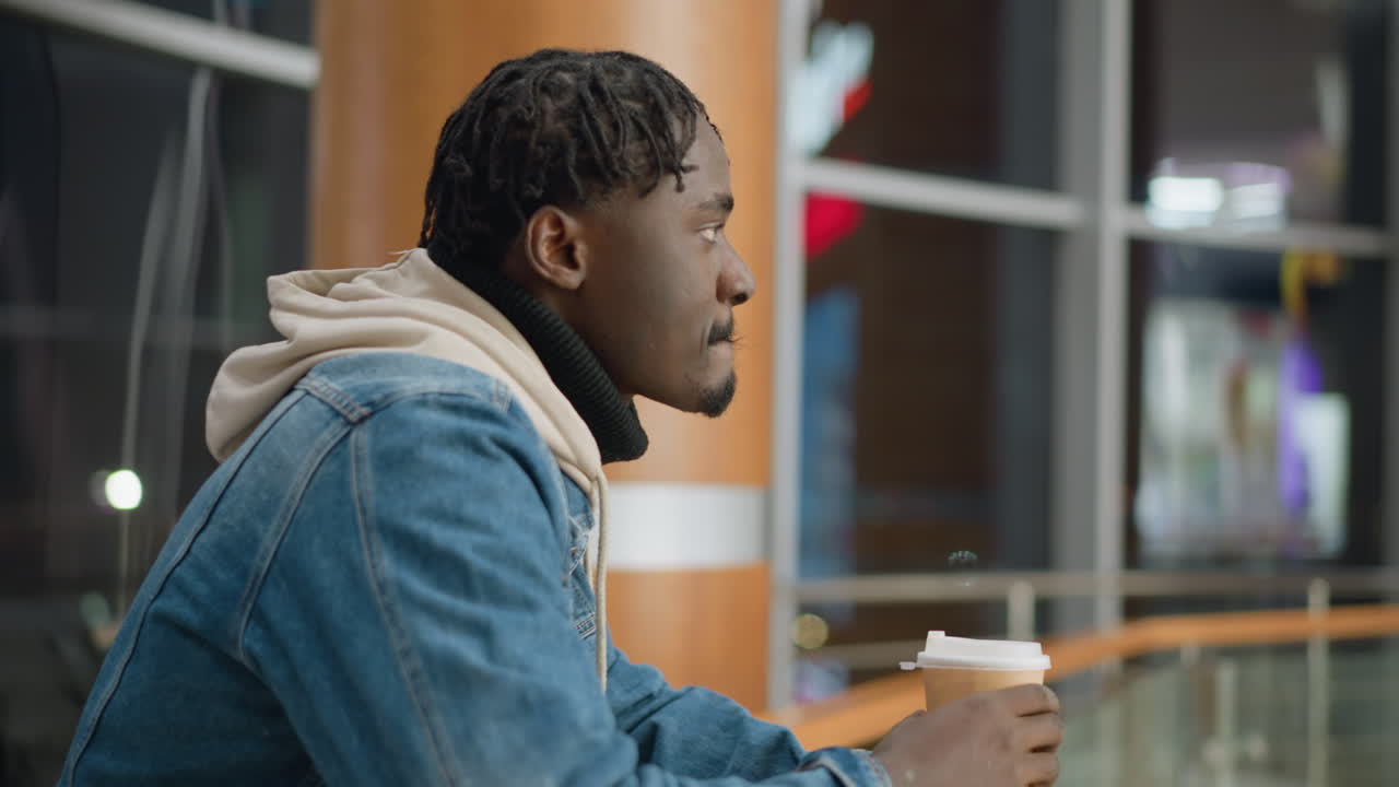 Young man in denim jacket and hoodie sipping from paper cup while resting on glass railing, looking contemplative and enjoying beverage in modern urban indoor space