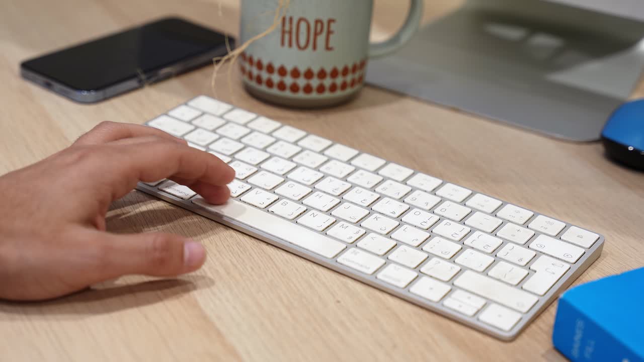 Primo piano di una mano che si allunga verso una tastiera bianca su una scrivania da ufficio in legno con smartphone, tazza di caffè e monitor del computer, mostrando una postazione di lavoro moderna e pulita