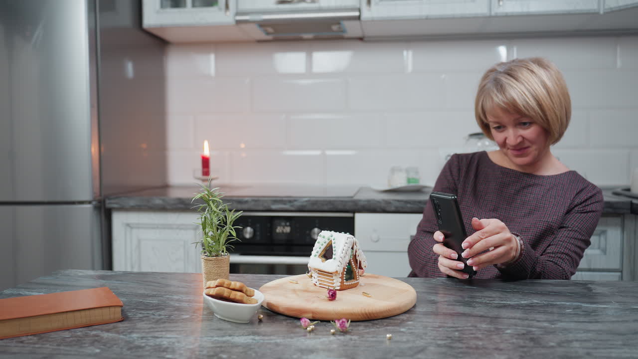 웃는 여성은 나무 트레이에 아이싱과 구슬을 넣은 아름답게 장식 된 진저브레드 집의 사진을 찍습니다. 부에는 불, 비 식물, 쿠키 및 시골 장식이 있습니다.