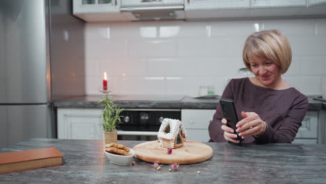 una mujer sonriente toma una foto de una casa de pan de jengibre bellamente decorada con hielo y cuentas en una bandeja de madera, la cocina cuenta con luz de vela, planta en olla, galletas y decoración rústica