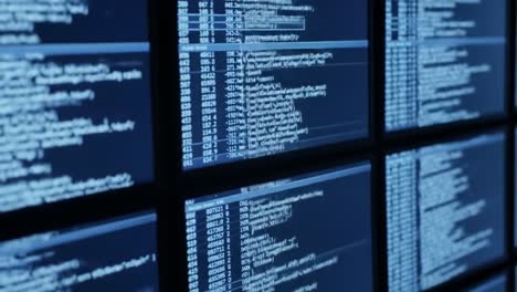 multiple computer screens display lines of code and data in a tech workspace at night. the setting reveals an atmosphere of focused programming and digital processing amidst a dark environment.