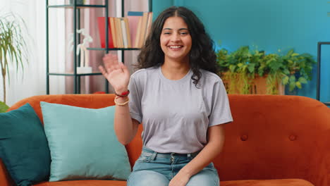 indian woman smiling friendly at camera, waving hands gesturing hello, hi, greeting at home on sofa