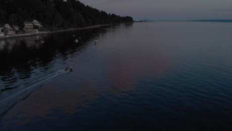 a twilight aerial view of a boat racing past parked boats floating in the water in front of a beach and homes on the shore