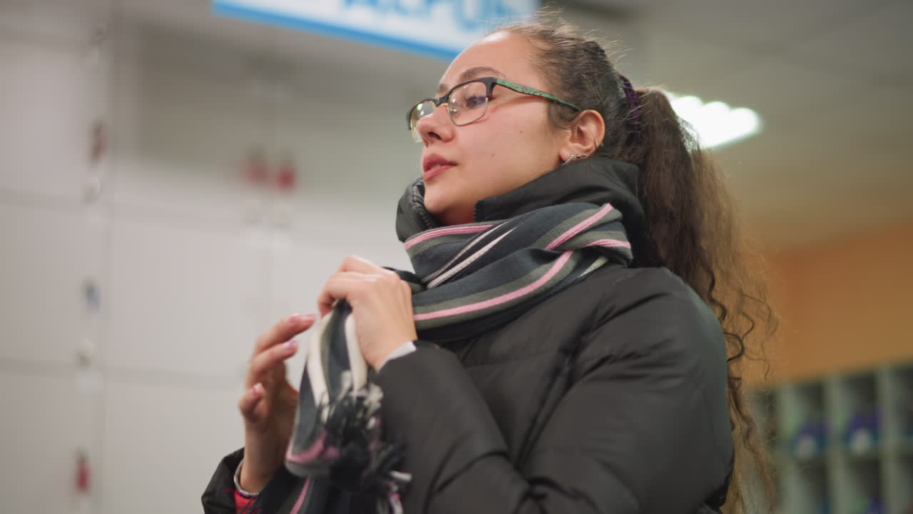 Damsel in glasses wearing black puffer jacket and striped scarf adjusting scarf indoors, calm expression, winter outfit, candid authentic lifestyle moment showing natural beauty in public environment