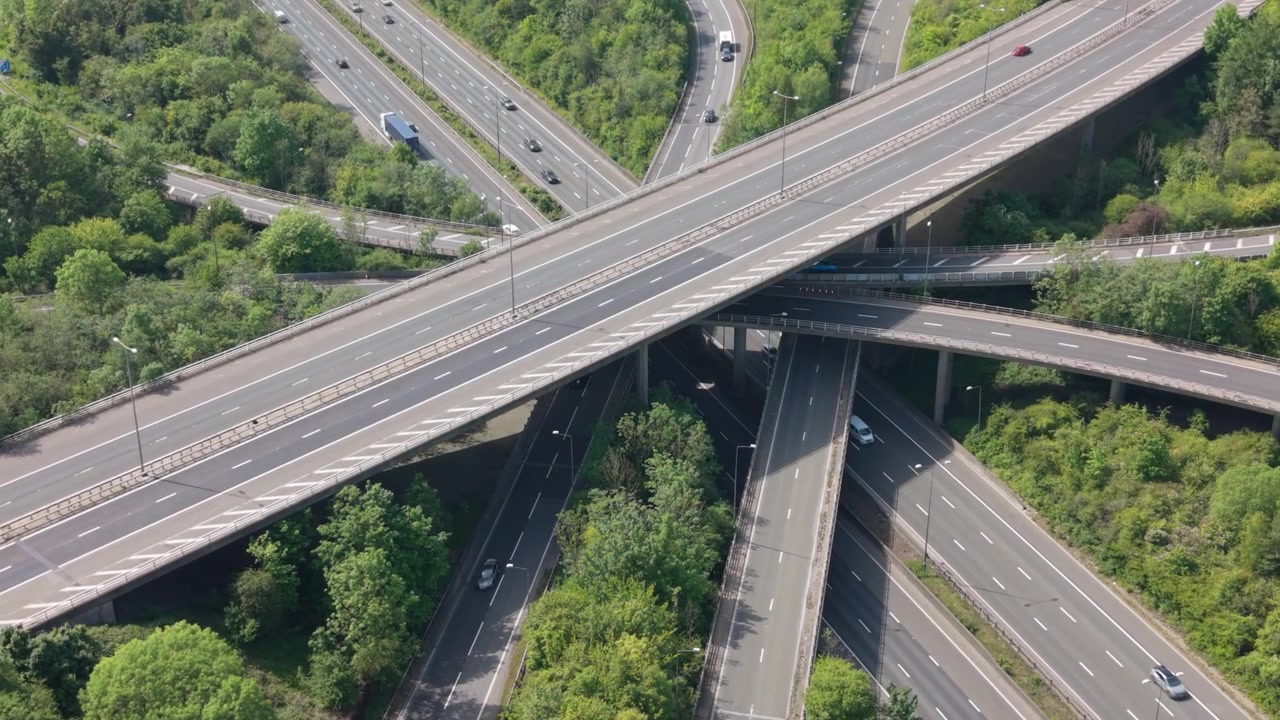 Una vista aérea captura el ajetreado tráfico diurno fluyendo a través de un complejo intercambiador de autopistas de varios niveles. Numerosos coches, camiones y furgonetas se mueven por las carreteras asfaltadas, rodeados de exuberantes árboles verdes.