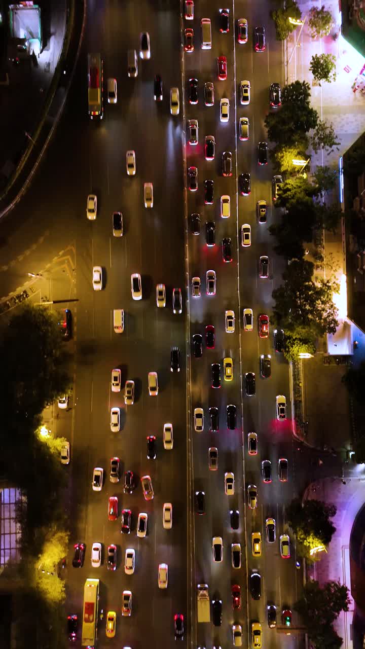 Vertical aerial shot of rush hour traffic in Guangzhou, China, at night. Cars and taxis move through the bustling city streets, illuminated by streetlights and surrounding buildings.