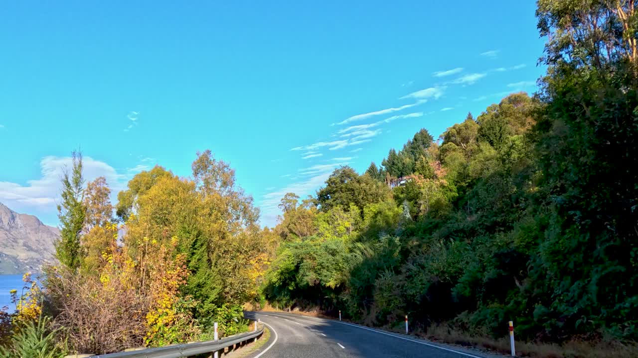 Vehicle travels winding country road through lush forest, mountains visible, bright daylight, smooth camera movement