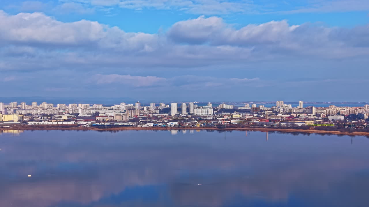 Bysilhuet langs floden med bygninger og skyer spejlet i vandet om dagen under en klar himmel