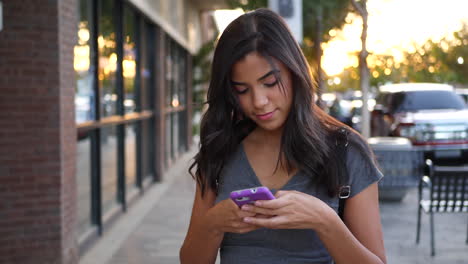 Una-Atractiva-Joven-Hispana-Caminando-Por-Las-Calles-De-La-Ciudad-Enviando-Mensajes-De-Texto-Y-Luciendo-Feliz-Con-Un-Mensaje-En-Su-Celular-A-Cámara-Lenta