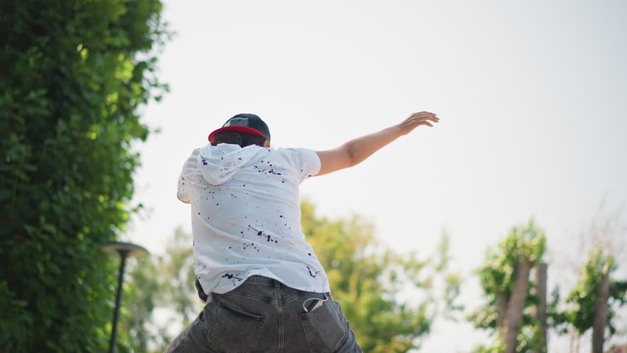 Skater dancing outdoors, Individual performing energetic dance moves beneath leafy trees in sunlight, Street dancer in cap and glasses engaging in lively movements beside park pathway in afternoon